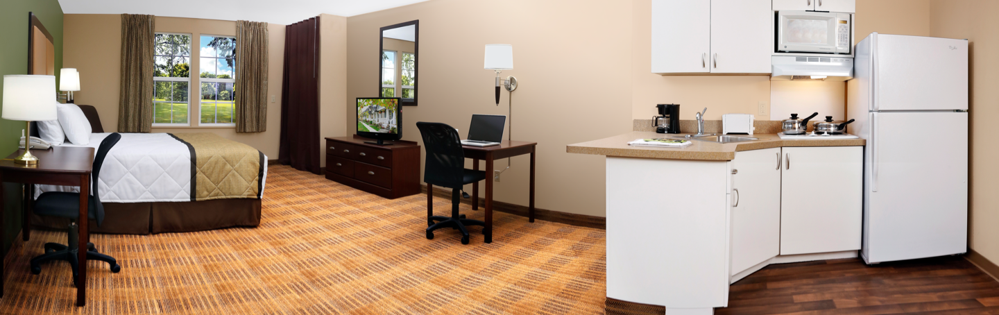 Panoramic view of a room with (from left to right) nightstand with lamp, bed, TV, desk and kitchen visible